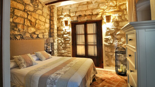A bedroom with a stone wall, wooden beams, a bed with a striped bedspread, a nightstand, a birdcage, and a dresser. - THE BALCONY OF THE OLIVE TREES (Luque)