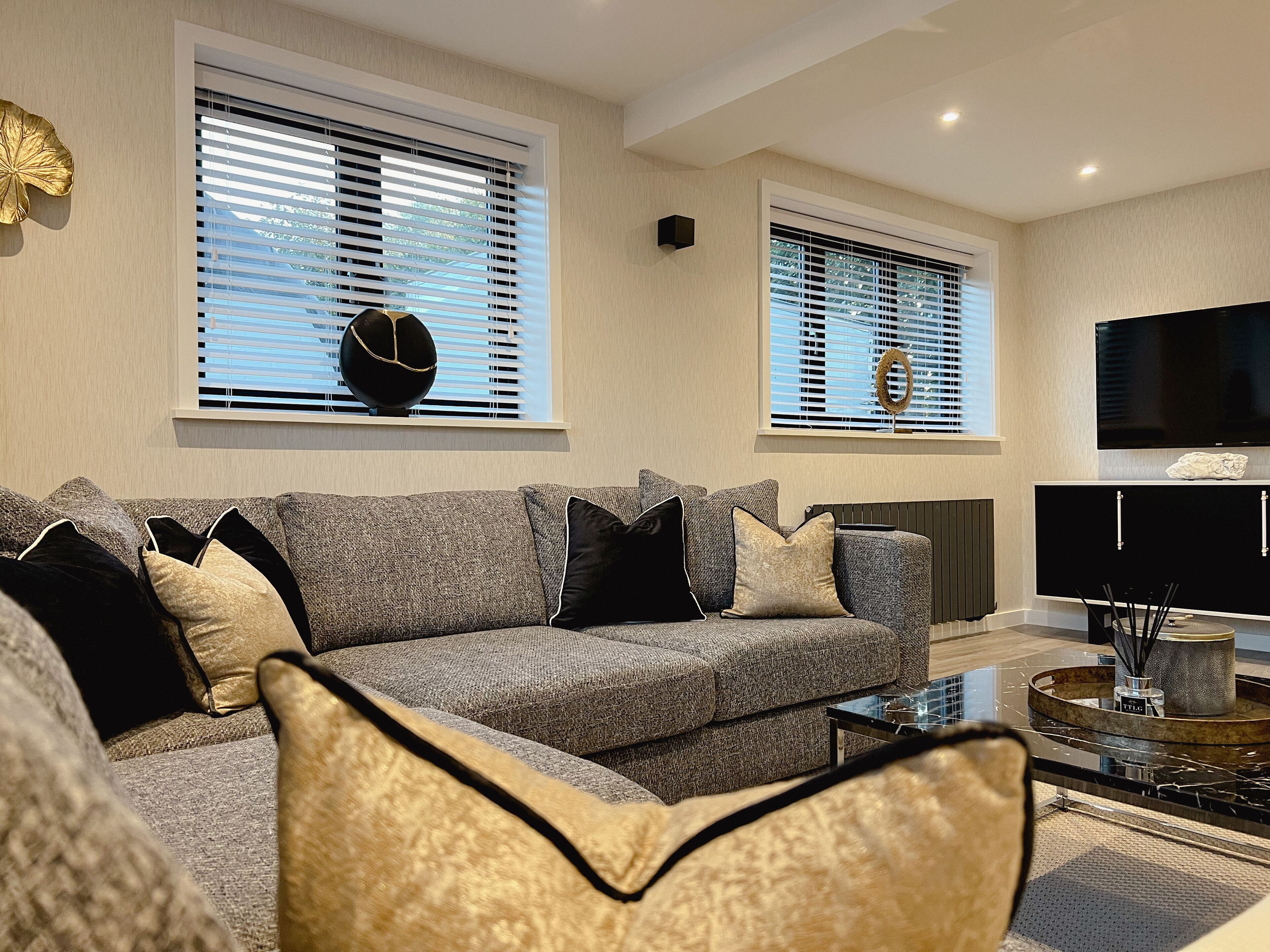 A modern living room with a grey sofa, black and gold cushions, a black coffee table, and a flat-screen TV mounted on the wall.