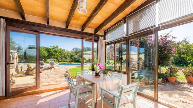 A sunlit patio with a table and chairs, large glass doors, and a view of a garden. - Pet-Friendly "Finca Pep" with Pool & Wi-Fi (Sineu)