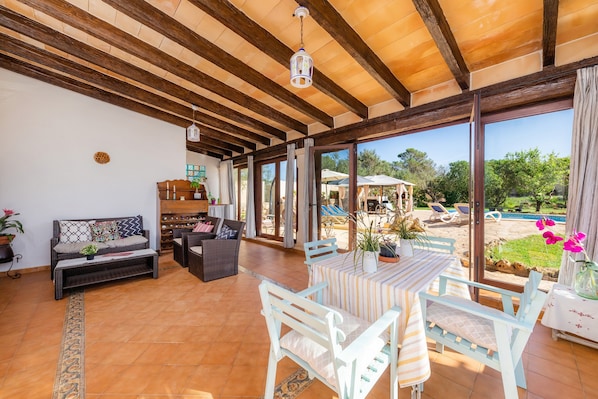 A spacious living area with a wooden ceiling, a sofa, a dining table with chairs, and a view of an outdoor swimming pool.