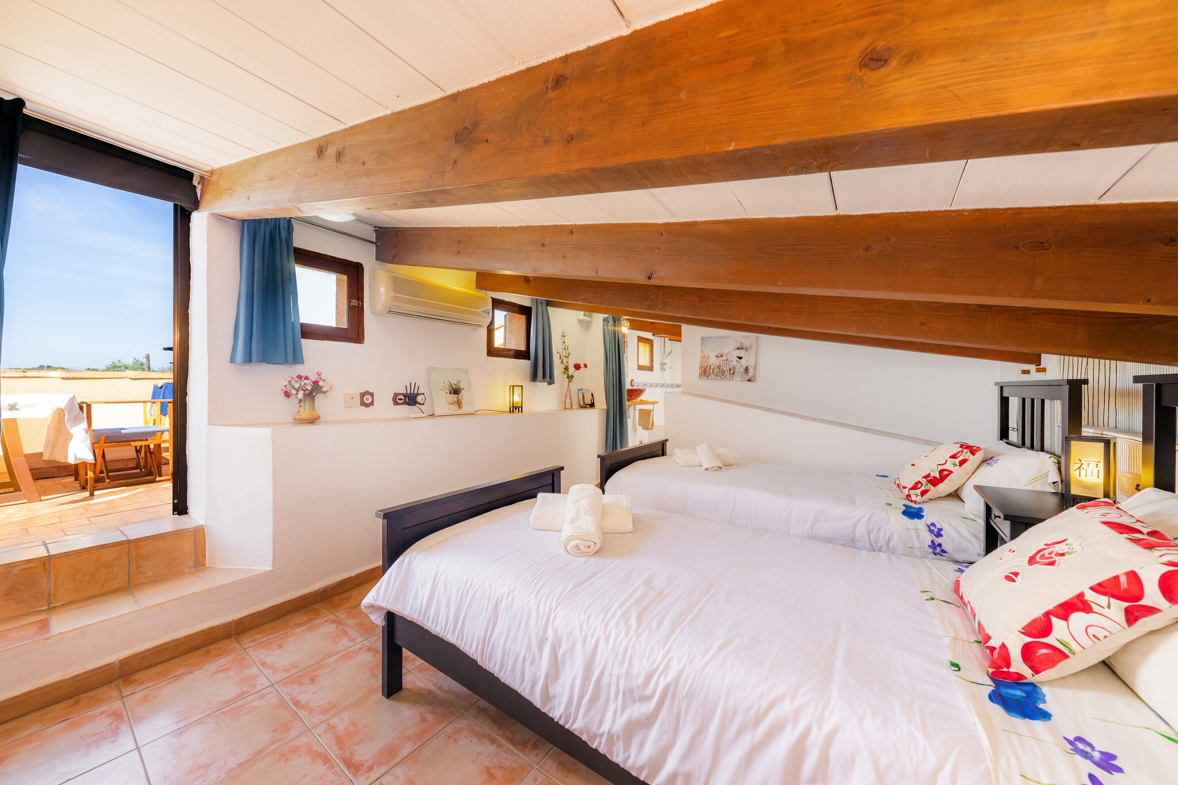 Ein Schlafzimmer mit einem großen Bett, einer Holzbalkendecke, einem kleinen Fenster mit Vorhängen und Blick ins Freie.