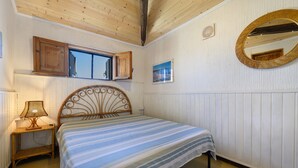 Une chambre à coucher comprenant un lit, une table de chevet, une lampe, une fenêtre avec un volet en bois, un miroir fixé au mur et un cadre photo.