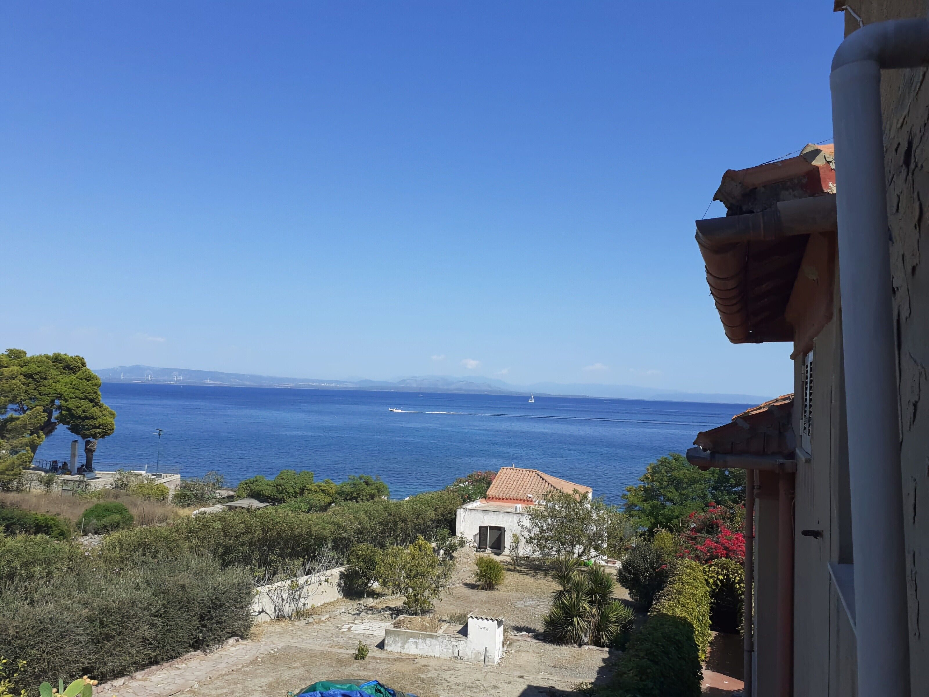 Blick auf die Küste mit blauem Himmel, ein kleines Gebäude mit rotem Ziegeldach und ein Gewässer mit einem Segelboot.