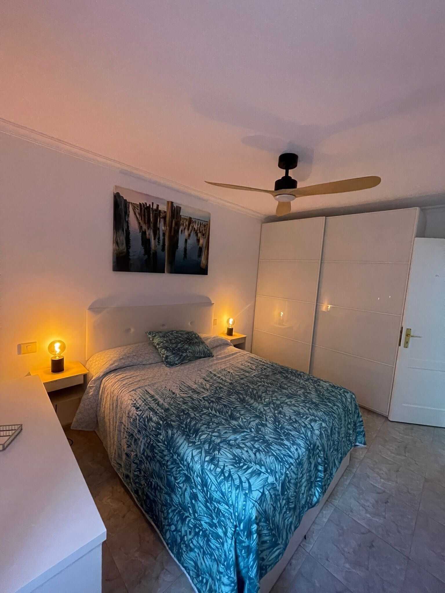 Een slaapkamer met een bed, twee nachtkastjes met lampen, een plafondventilator, een kledingkast en een schilderij aan de muur.