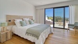 Een slaapkamer met een groot bed, een nachtkastje, een lamp en uitzicht op een balkon met buitenmeubilair.