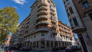Un immeuble à plusieurs étages doté d’un balcon d’angle, de boutiques au rez-de-chaussée et de voitures garées devant.