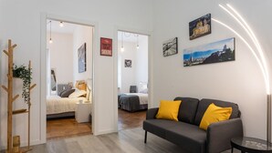 Un salon moderne avec un canapé, une table basse et un porte-manteau en bois. On aperçoit deux lits au fond de la pièce, l’un recouvert d’une couette blanche et l’autre d’une couette grise.