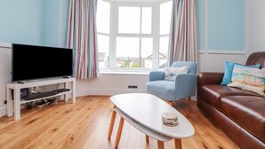 Una sala de estar con una televisión, una mesa de centro blanca, un sofá de cuero marrón y un sillón azul.