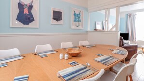 Comedor con una mesa redonda dispuesta para seis personas, sillas blancas, obra de arte enmarcada en la pared y un televisor al fondo.