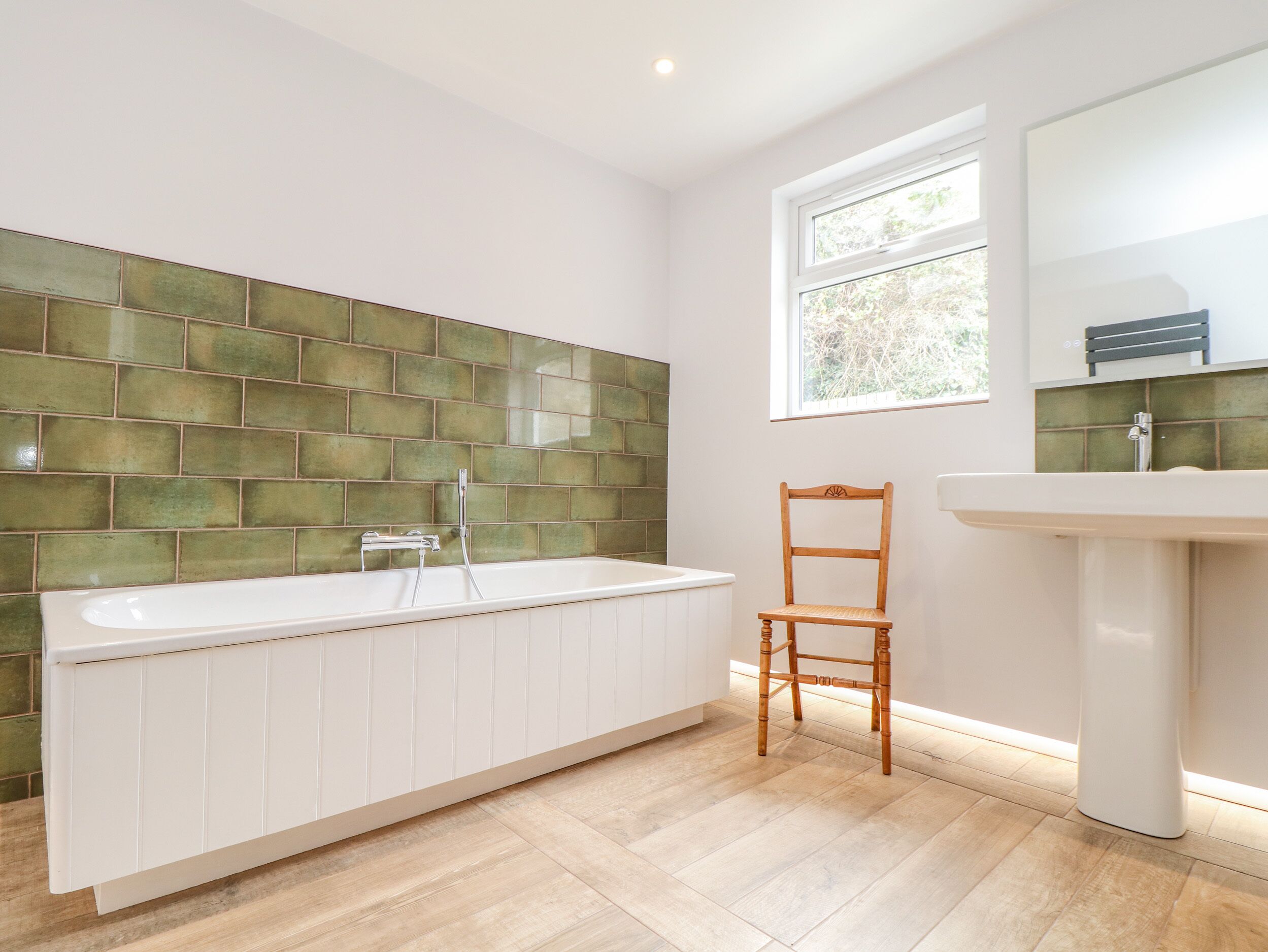 A bathroom with a white bathtub, a wooden chair, a white sink, and a window with a view of greenery.