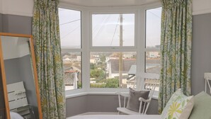 Un dormitorio con un ventanal grande, una cama con sábanas blancas, un armario de madera y un sillón con cojines.