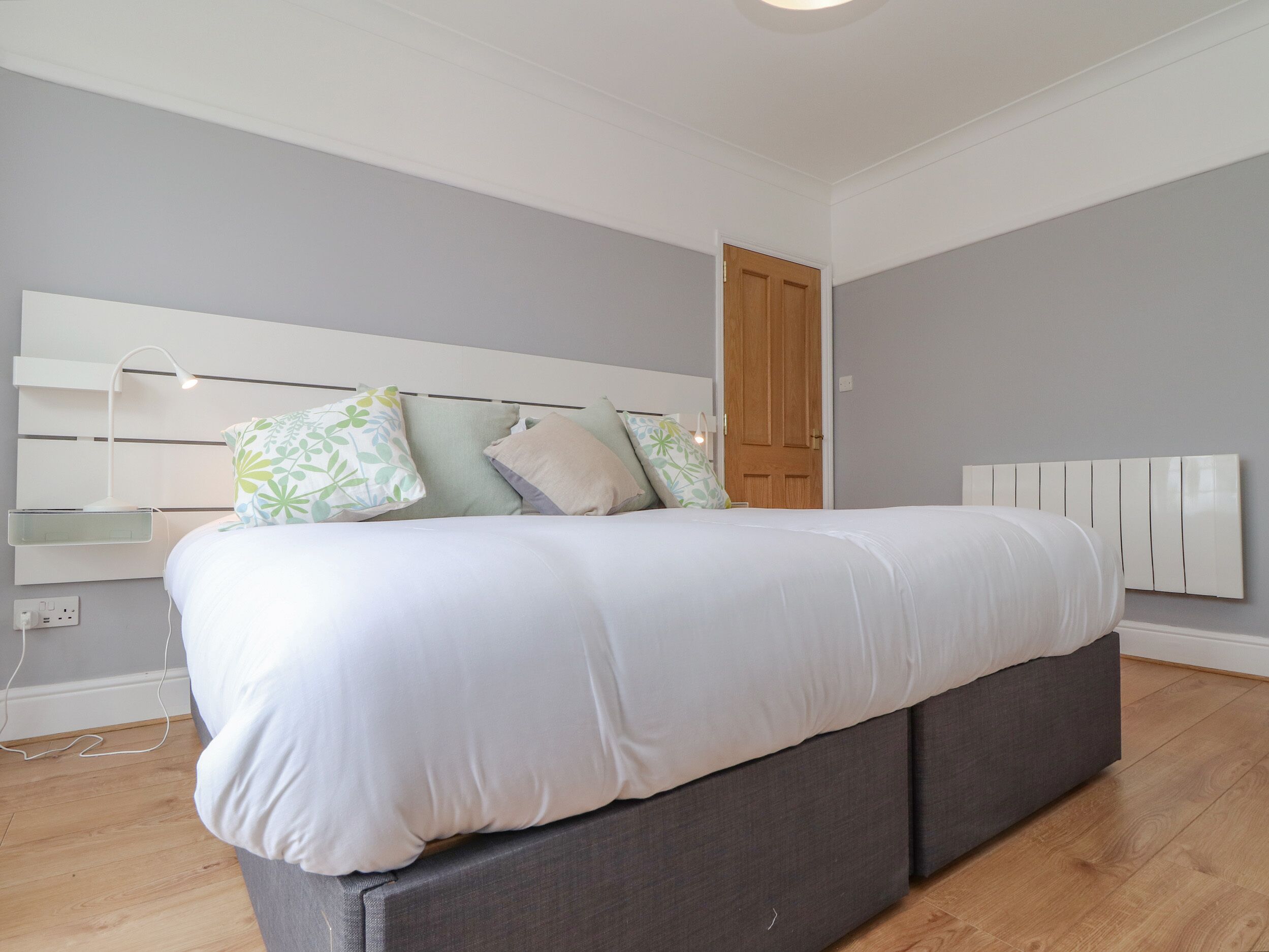 A modern bedroom with a grey upholstered bed, a white headboard, and a wooden door.