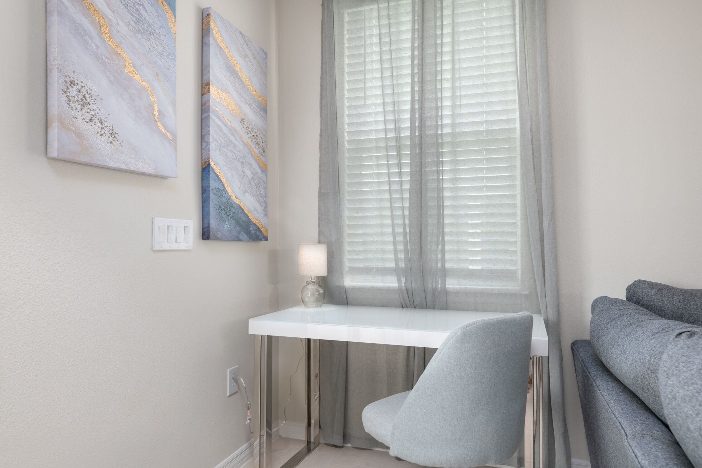 A modern room with a white desk, a grey chair, and a grey sofa. There are two abstract paintings on the wall and a window with blinds.