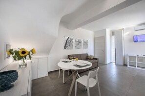 Uma sala de estar moderna e bem iluminada, com uma mesa de jantar redonda, cadeiras brancas, um sofá e uma TV fixada na parede.