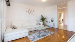 A modern living room with a white sofa, a glass coffee table, and a wooden floor. There is a decorative wall piece above the sofa and a plant on the right side. - Apartment Parentium - Stay in the Heart of Porec (Porec)