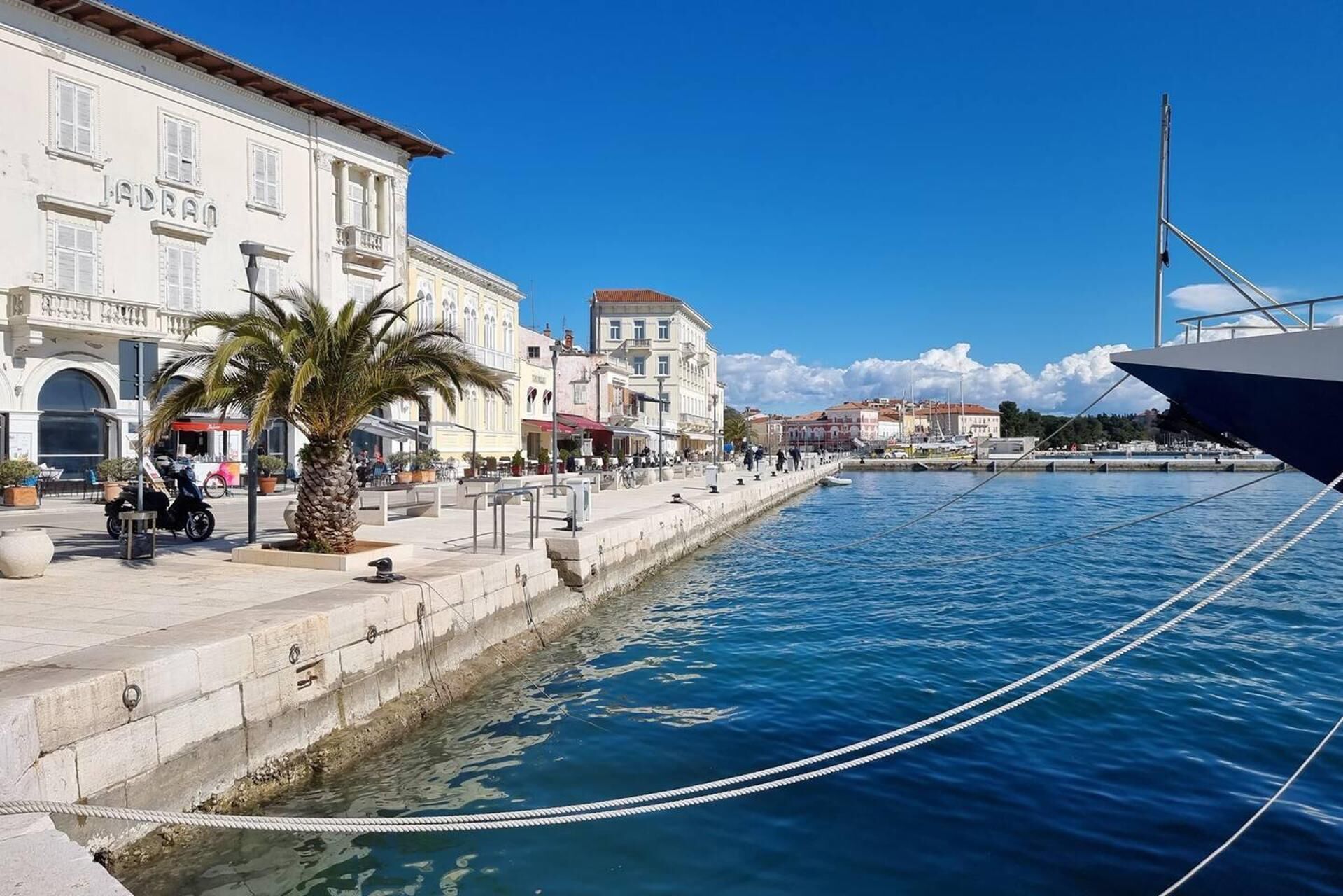 A waterfront promenade with a building labeled 'JADRAQ' and a large yacht docked nearby.