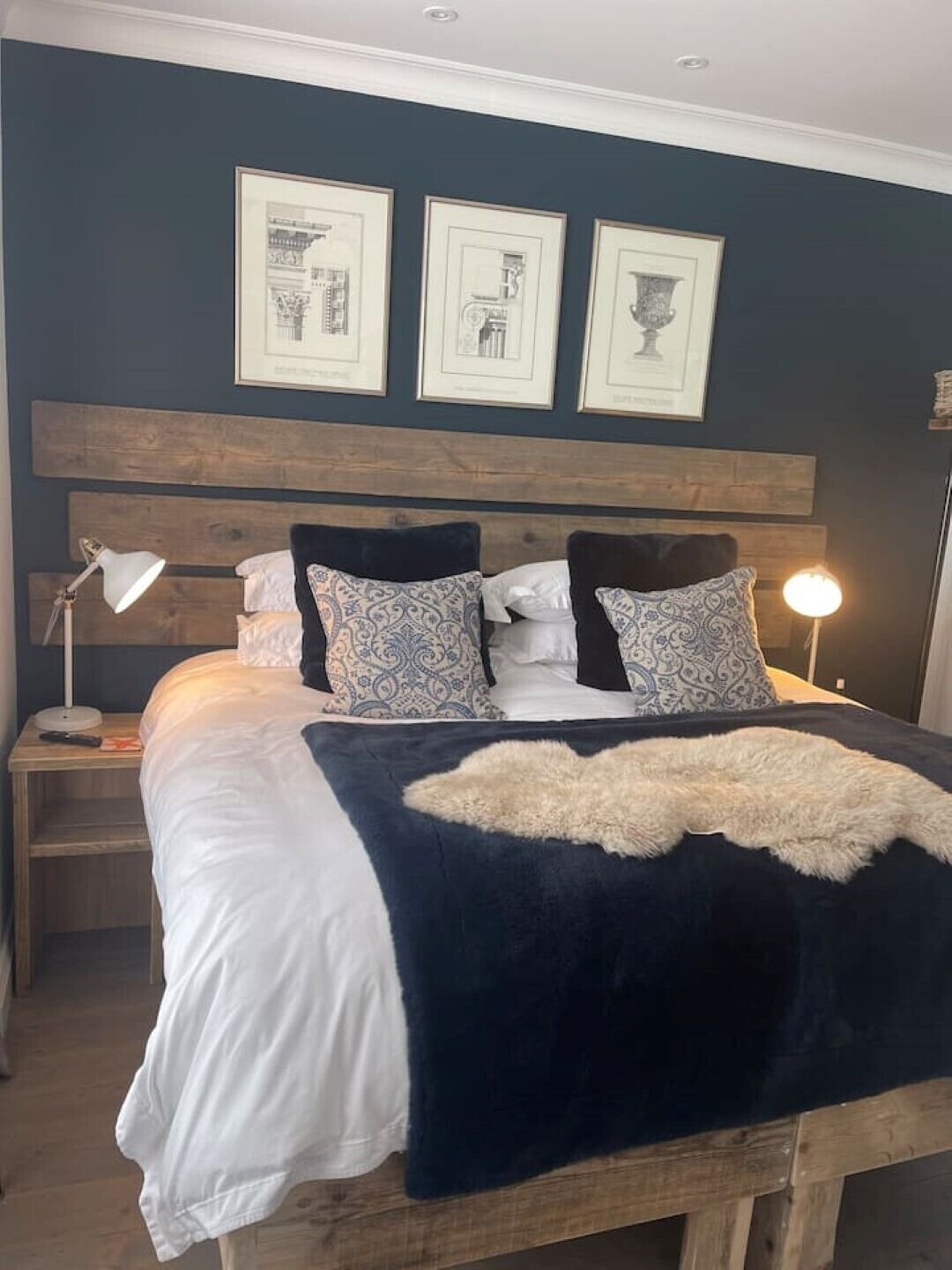 Una camera da letto con testiera in legno, un letto grande con biancheria da letto bianca e blu scuro, cuscini decorati, una coperta soffice e disegni architettonici incorniciati appesi al muro.