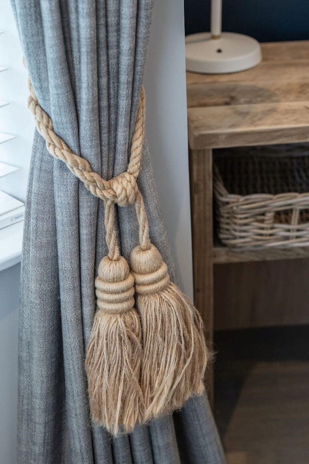 Een macramé wandversiering met kwastjes voor een grijze gordijn.