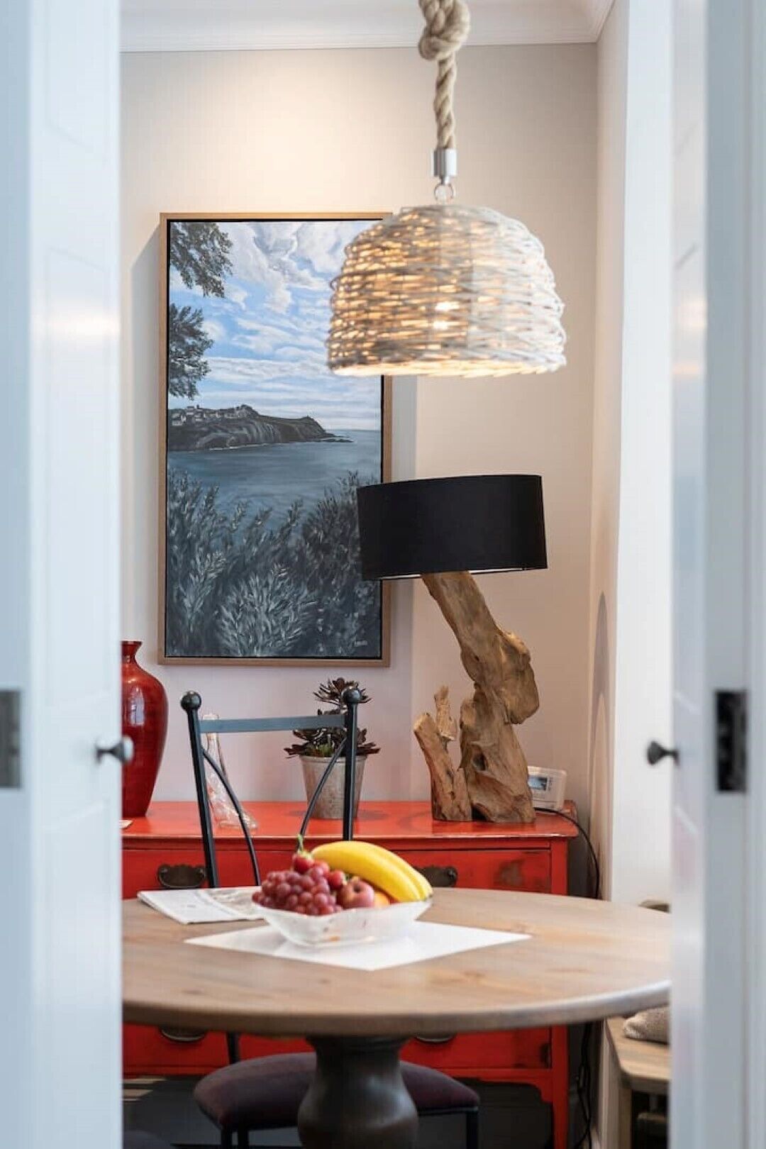 Un comedor con una mesa redonda, una silla roja, un frutero, una lámpara negra y un cuadro en la pared.