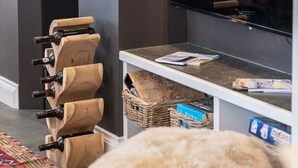 Ett modernt vardagsrum med en platt-TV som är monterad på väggen, ett träställ för vinflaskor, ett litet bord med tidningar och ett fönster med persienner.