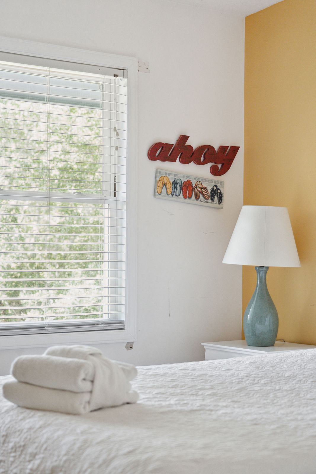 A hotel room with a bed, a window with blinds, a wall-mounted sign reading "ahoy", a lamp, and a towel on the bed.
