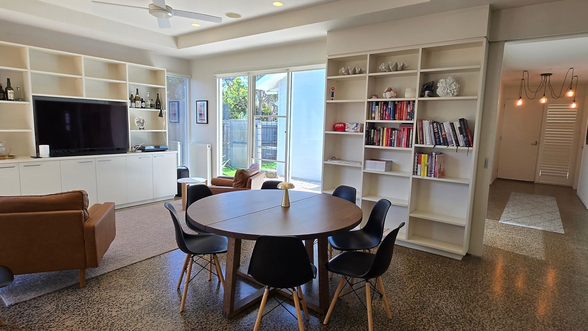 A modern living room with a round dining table, black chairs, a brown sofa, and a TV on a white cabinet. There are bookshelves on both sides of the room, and a large window with a view of the outdoors.