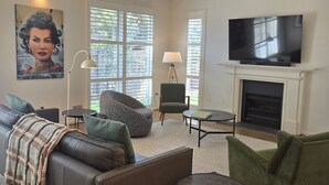 Un salon avec un canapé en cuir, un fauteuil vert, un pouf, une petite table, une lampe, un tableau, une cheminée et un téléviseur.