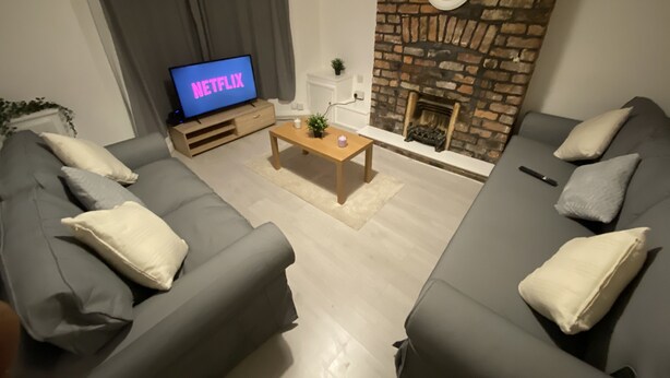 Moderni olohuone, jossa on harmaat sohvat, puinen sohvapöytä, televisio, jossa näkyy teksti »NETFLIX», ja tiilistä tehty takka.