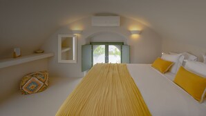 A bedroom with a bed, a bedside table, a window, and an air conditioning unit. - Two-bedroom Family Cave Suite with private terrace, kitchenette | Element Suites (Thira - Santorini)