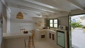 A compact kitchen with a white countertop, wooden stools, a hanging light fixture, a built-in microwave, and a window with green shutters. - Two-bedroom Family Cave Suite with private terrace, kitchenette | Element Suites (Thira - Santorini)