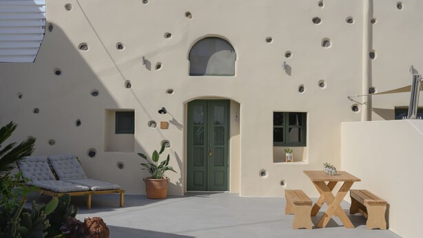 A modern outdoor patio with a cushioned lounge chair, a wooden table with benches, potted plants, and a green door. - Two-bedroom Family Cave Suite with private terrace, kitchenette | Element Suites (Thira - Santorini)