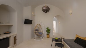 A modern living room with a sofa, a hanging chair, a small table, and a television mounted on the wall. - Deluxe Cave Suite with large private terrace & kitchenette | Element Suites (Thira - Santorini)