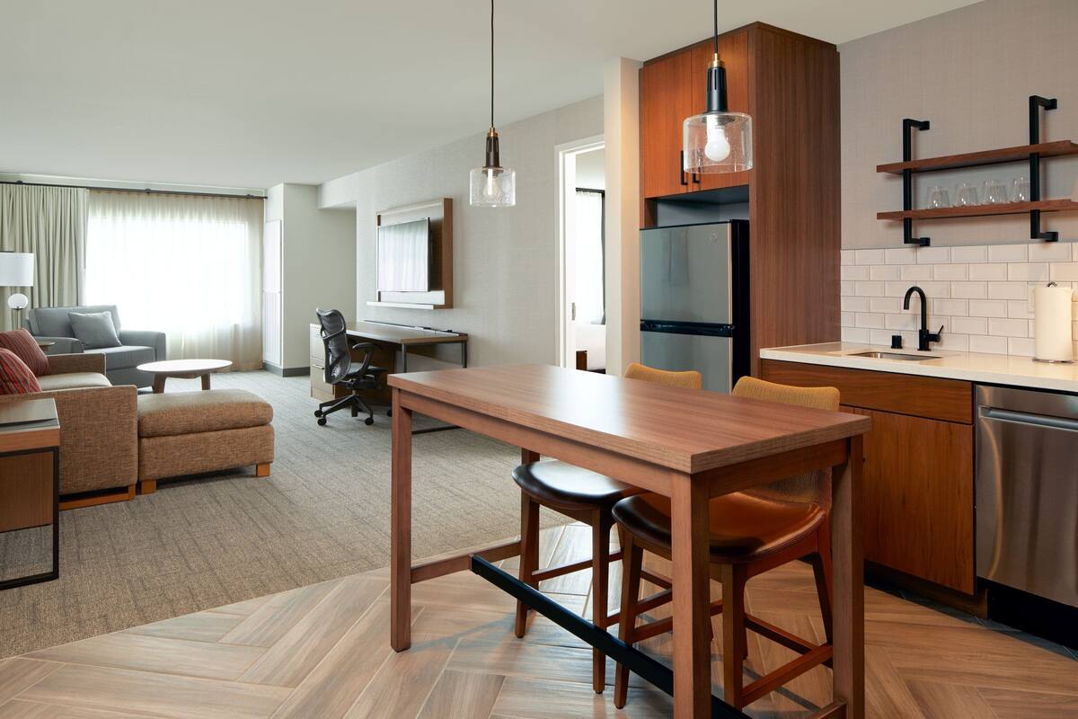 A modern hotel room with a wooden dining table, bar stools, a kitchen area with a refrigerator and dishwasher, a sofa, a chair, and a desk.