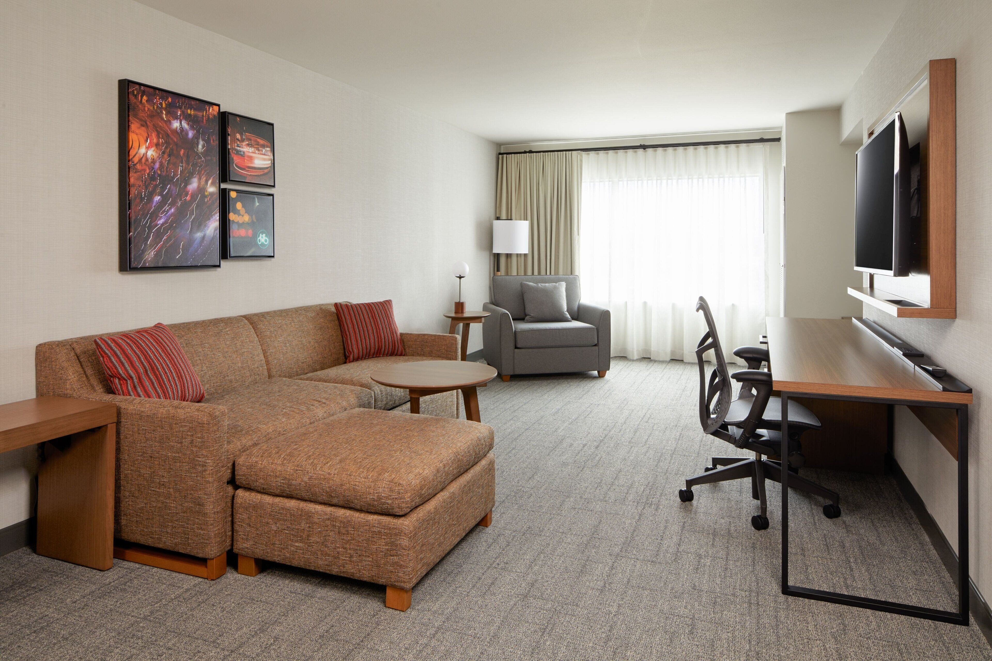Een moderne hotelkamer met een hoekbank, een klein rond tafeltje, een bureau met stoel, een aan de muur bevestigde televisie en kunstwerken aan de wand.