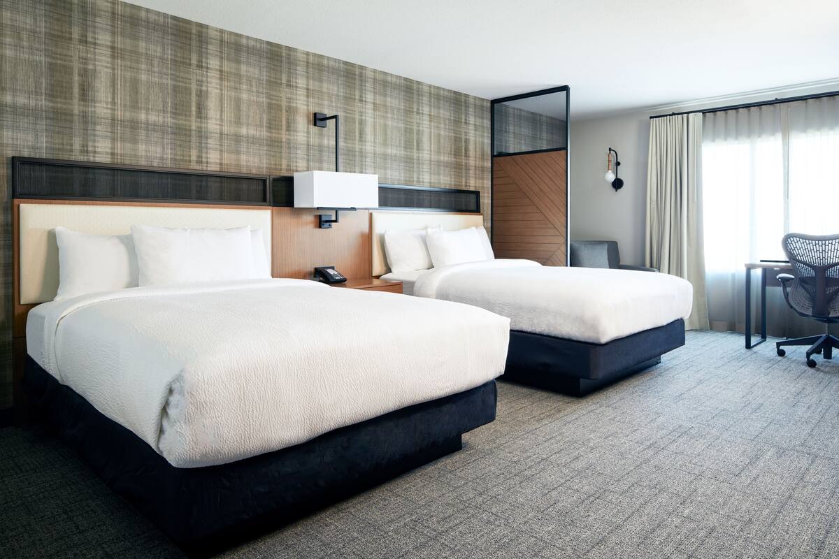 A hotel room with two beds, a desk, a chair, and a large window with curtains.