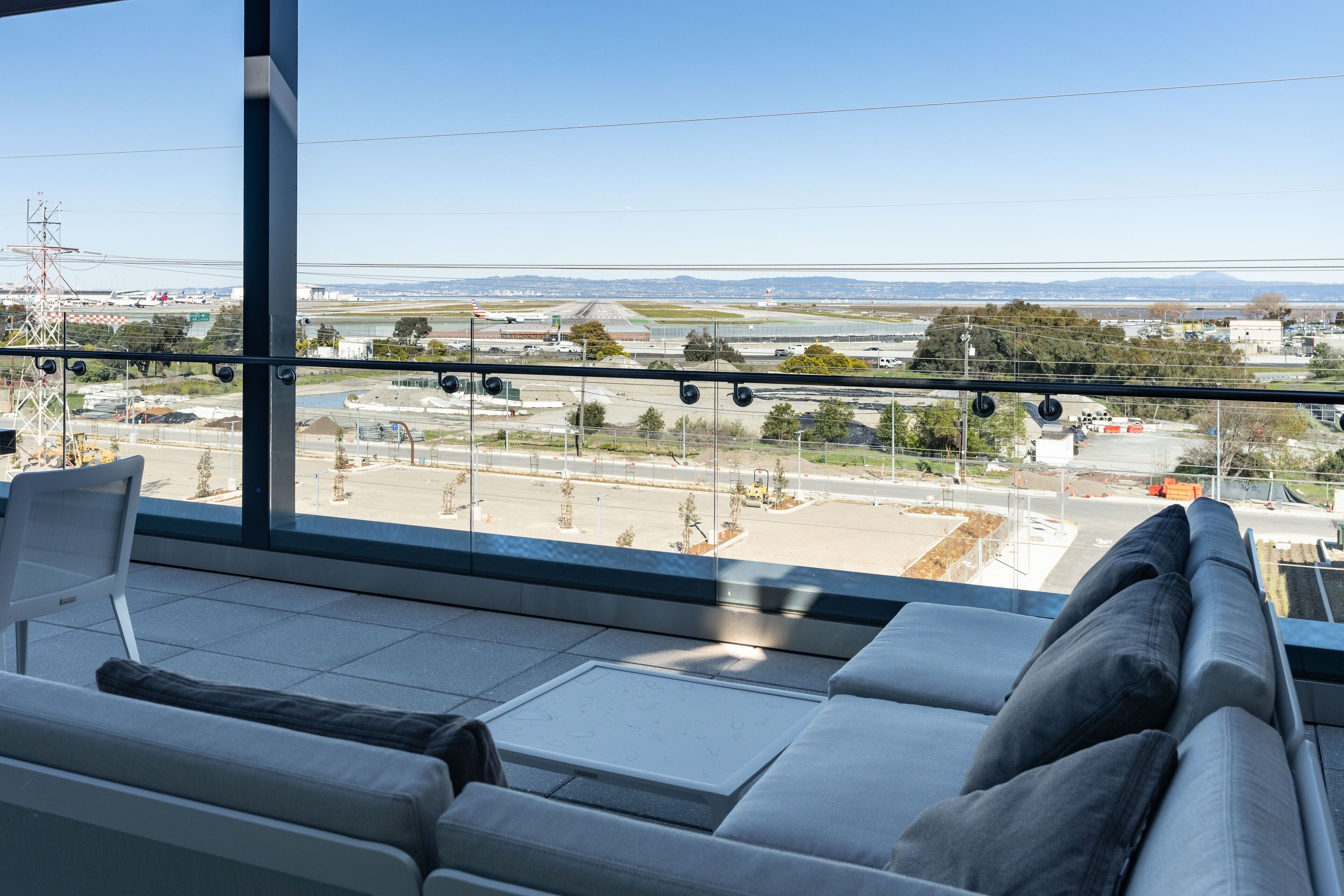A modern hotel room with a large window offering a view of an airport and distant mountains.