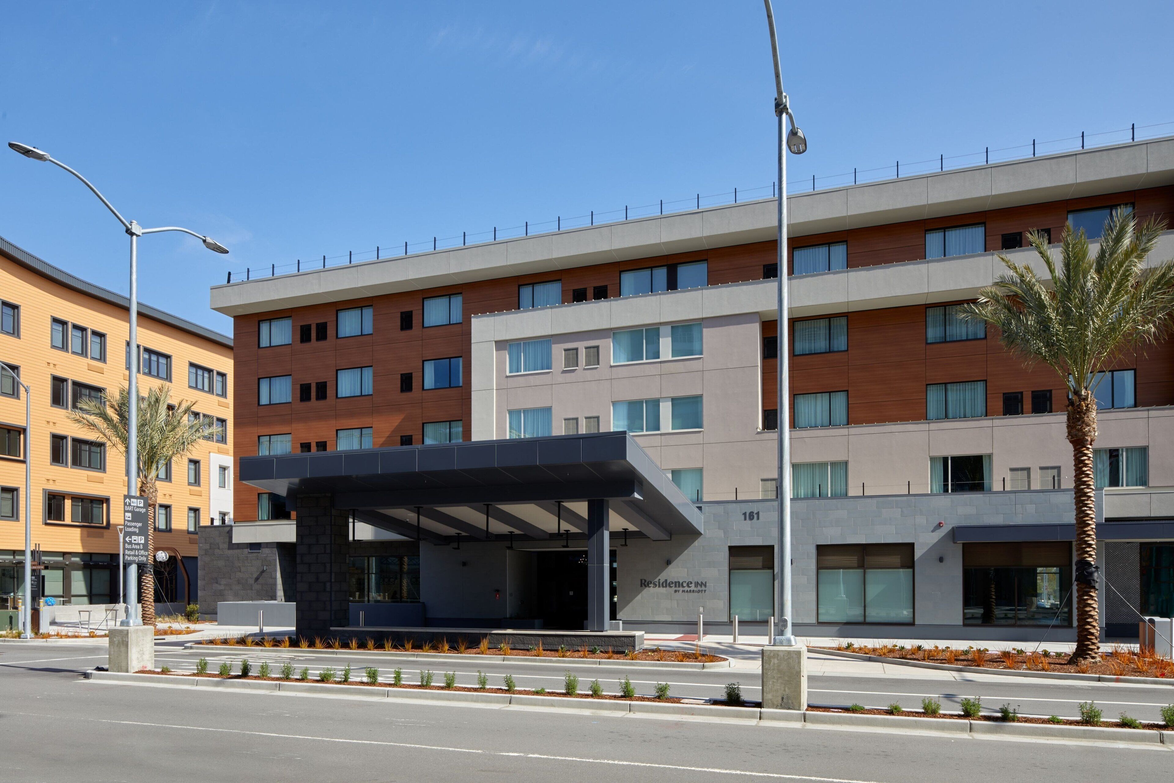 A modern hotel with a covered entrance, multiple floors, and a palm tree in the foreground.
