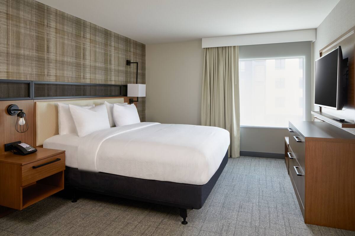 A hotel room with a large bed, a bedside table with a phone, a TV, and a window with curtains.