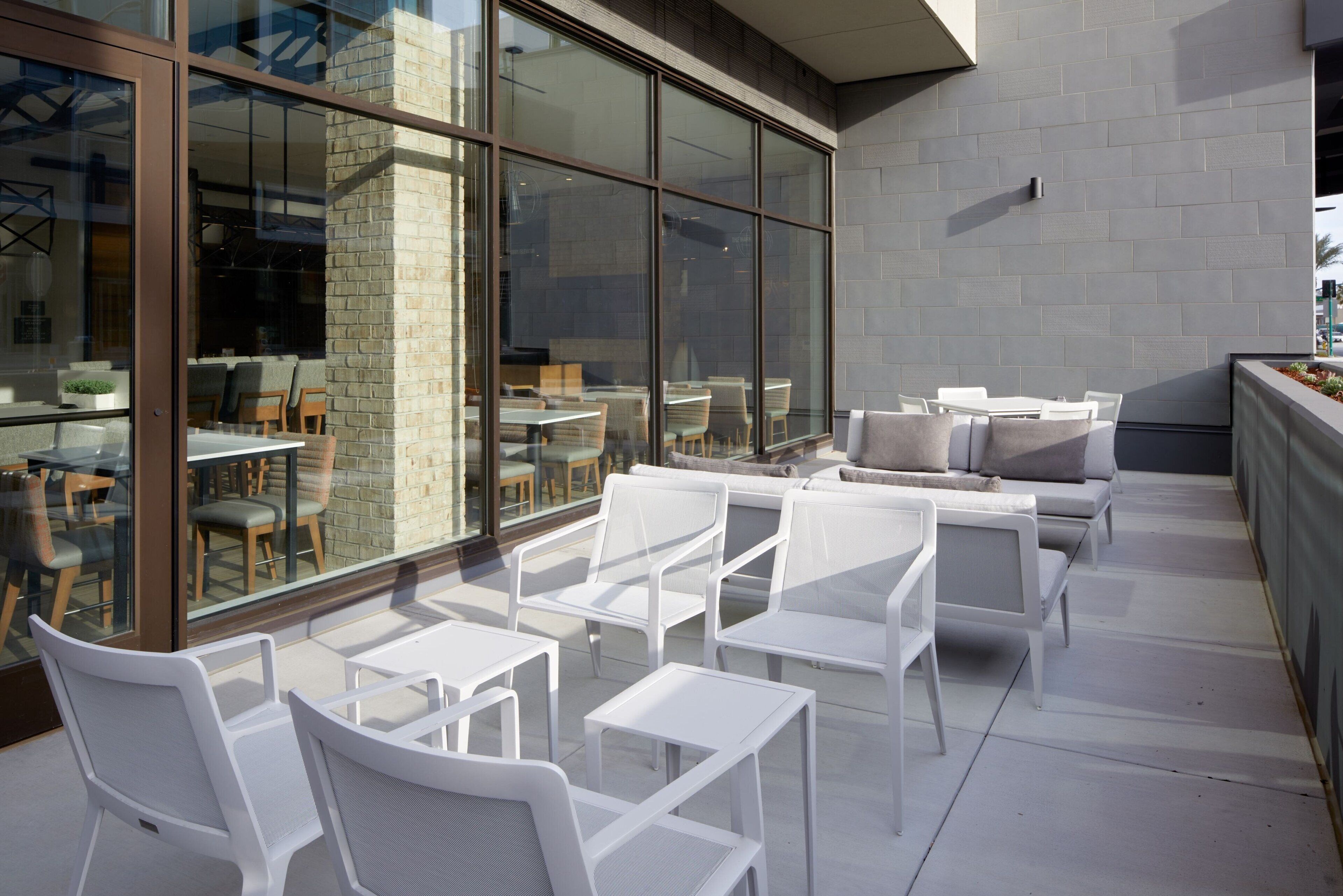 An outdoor seating area with white chairs and tables, large windows, and a brick wall.