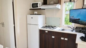 A compact kitchen with a refrigerator, microwave, gas stove, and a window with a view of greenery. - Location de Mobil Home Camping Tohapi en Alsace (Biesheim)