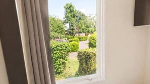 A bedroom with a bed, a window with curtains, a TV on the wall, and a view of a garden. - Location de Mobil Home Camping Tohapi en Alsace (Biesheim)