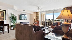 A modern hotel room with a living area featuring a sofa, armchairs, a coffee table, and a TV. There is a balcony with a view of the city. - Yacht Club Villas #2-405 Extraordinary and Spacious Condo! (North Myrtle Beach)