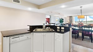 A modern kitchen with white cabinets, a black countertop, and a dishwasher. There is a dining area with a table and chairs visible in the background. - Yacht Club Villas #2-405 Extraordinary and Spacious Condo! (North Myrtle Beach)