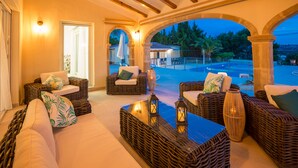 A cozy outdoor seating area with wicker furniture and cushions, a glass table, and a view of the pool and trees.