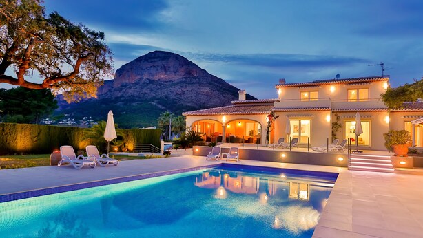A poolside area with lounge chairs, a swimming pool, and a house with a mountain in the background.