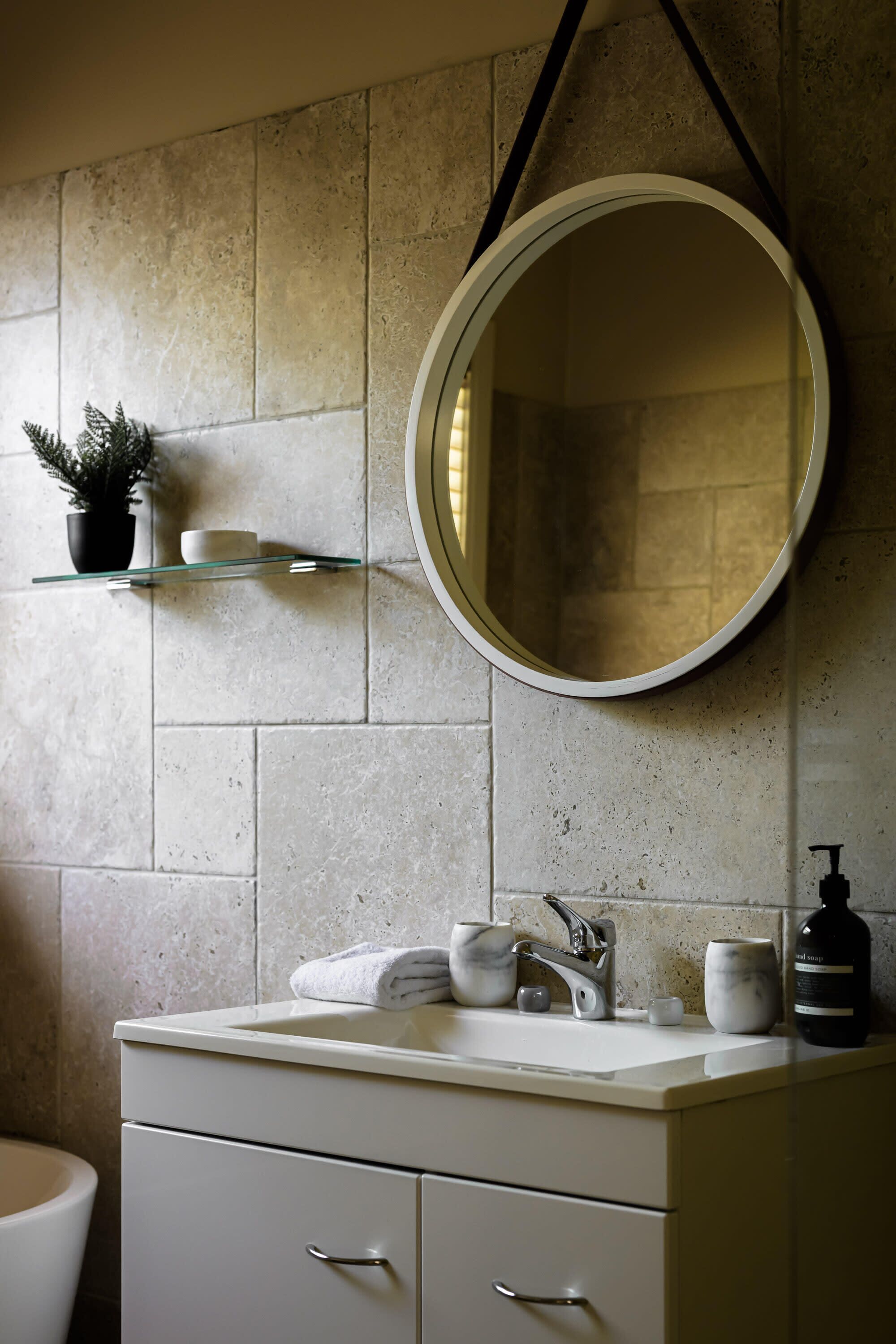 A bathroom with a white vanity, a round mirror, a towel rack, and a soap dispenser.
