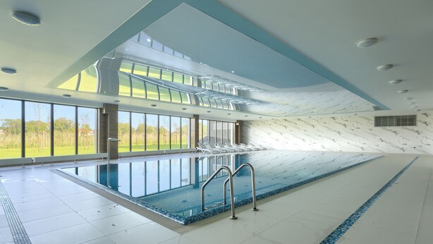 Una amplia piscina interior con paredes de vidrio transparente, suelo de baldosas y zona de la piscina con tumbonas.