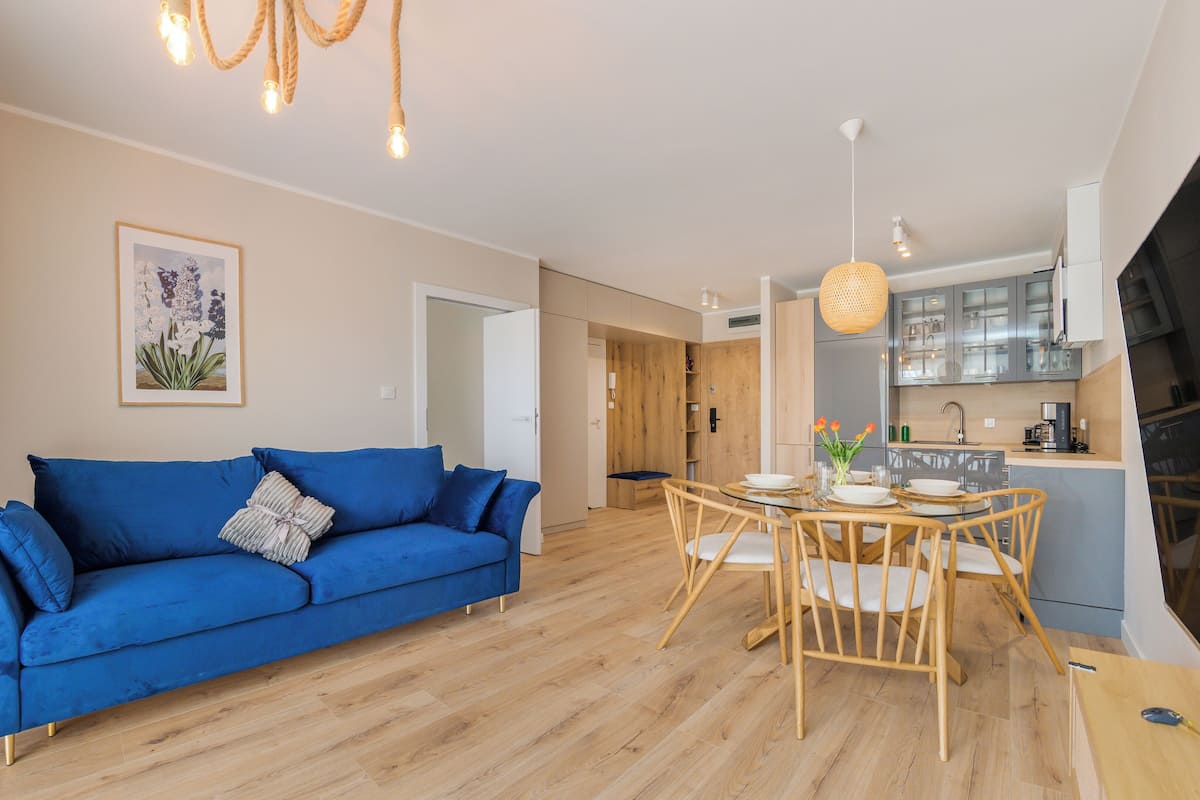 A modern living room with a blue sofa, wooden dining table, and a kitchen area with cabinets and appliances.