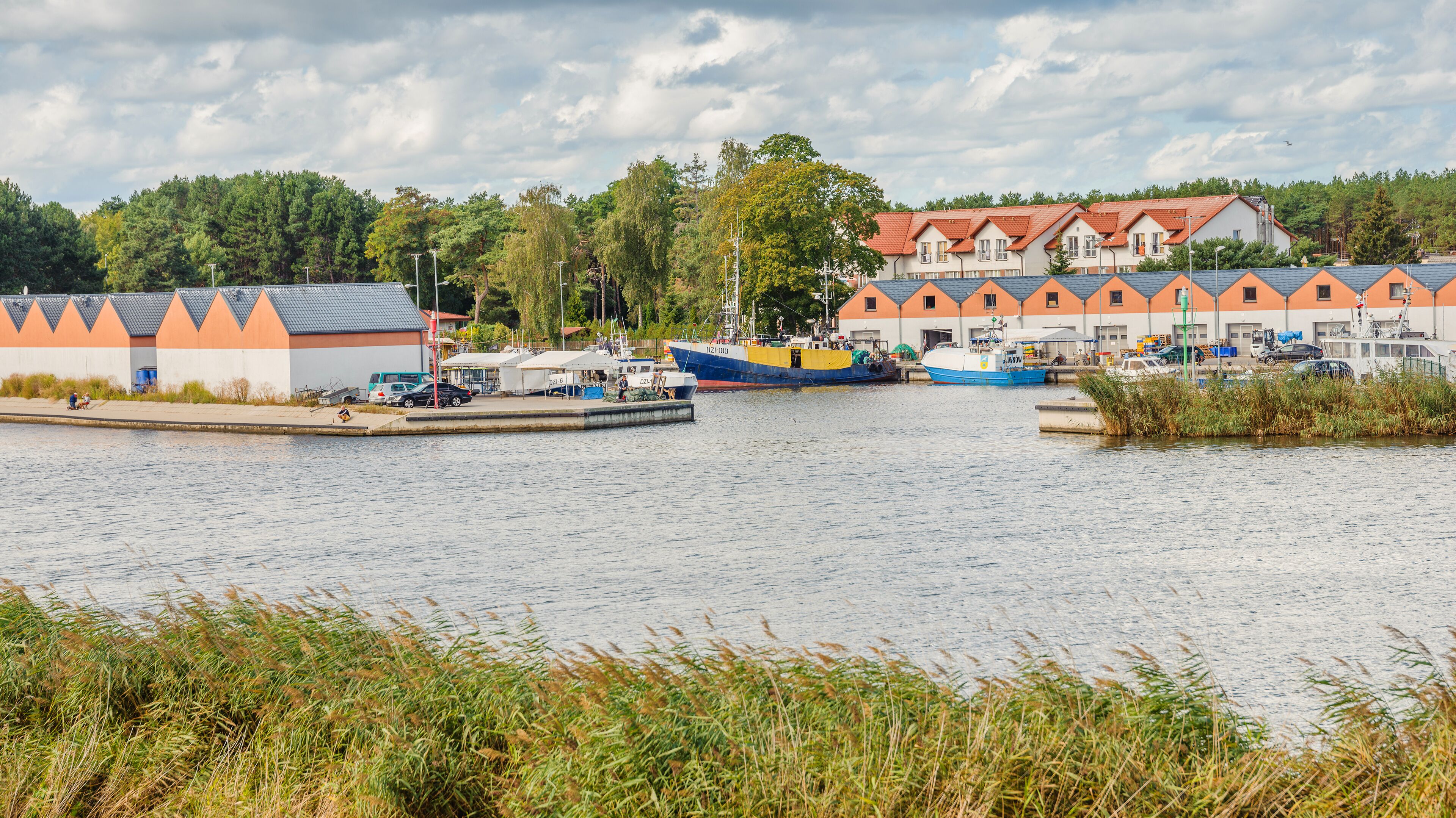 Un quai au bord de la rivière où sont amarrés des bateaux, une rangée de bâtiments aux toits orange et une colline boisée.