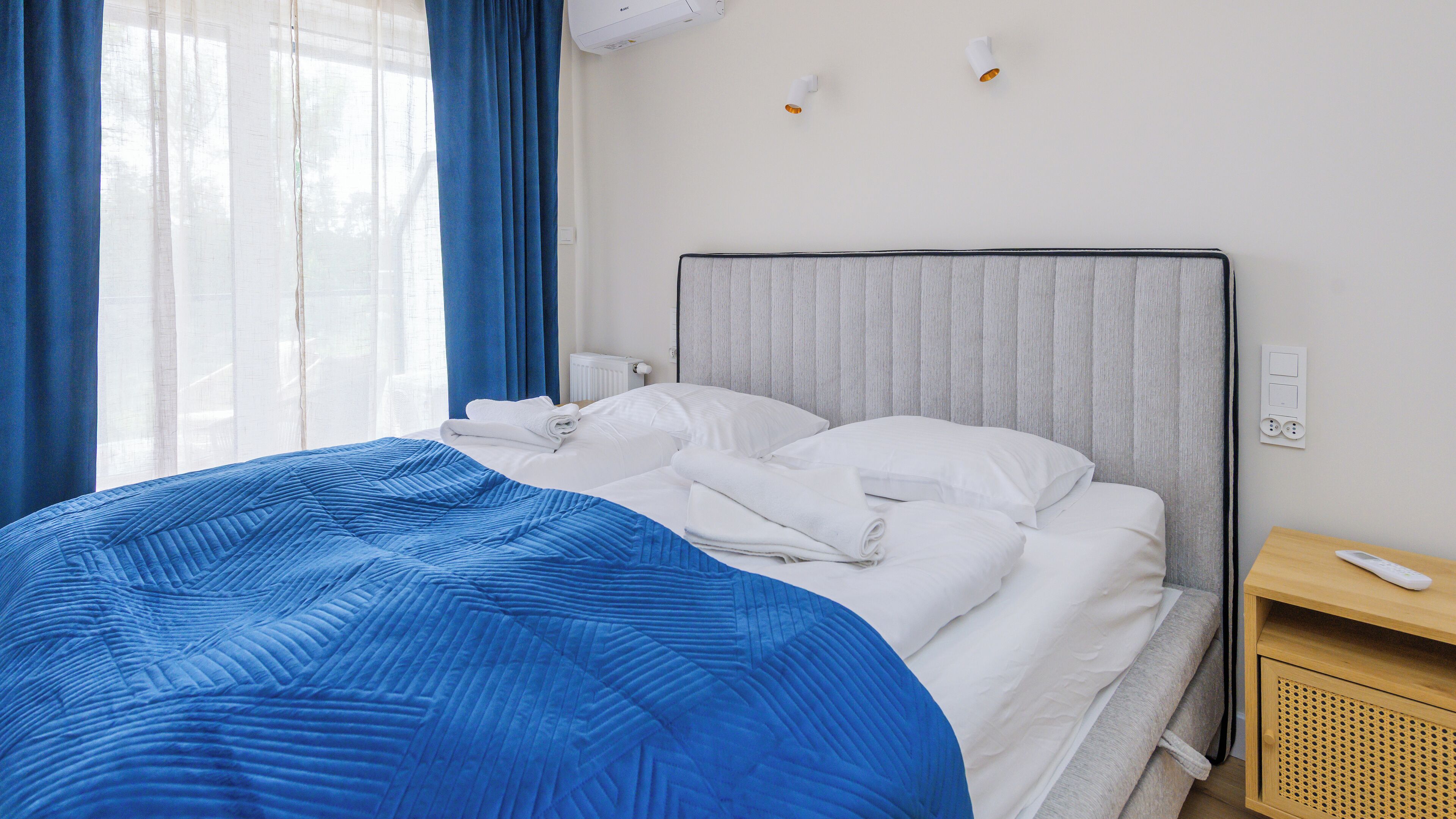 Un lit bien fait, avec des draps blancs et une couverture bleue, une table de chevet avec une télécommande et une fenêtre ornée de rideaux bleus.